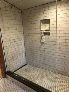 A bathroom with white tiles and a walk in shower with a glass door.