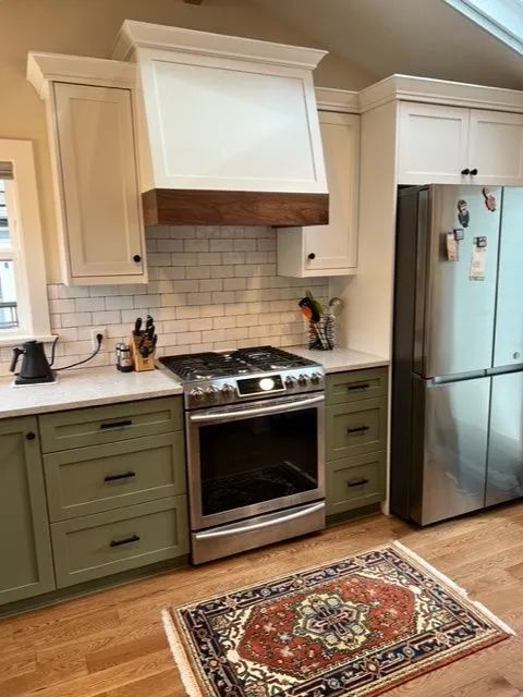A kitchen with green cabinets, a stove, a refrigerator and a rug on the floor.
