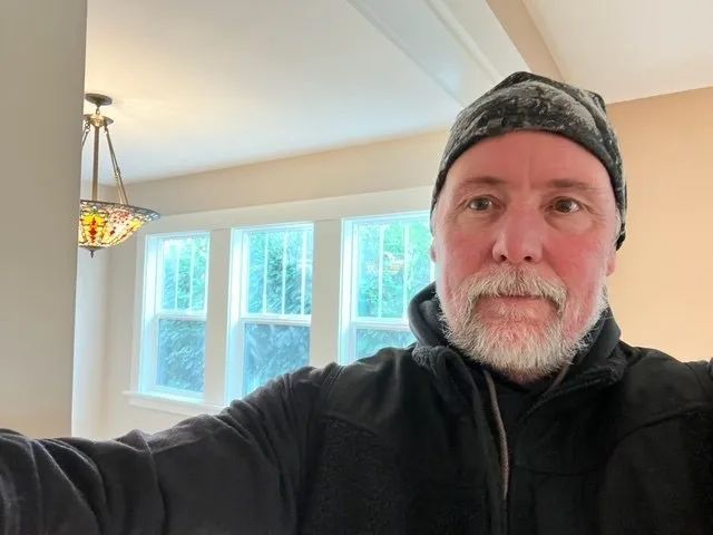 A man with a beard is taking a selfie in a living room.