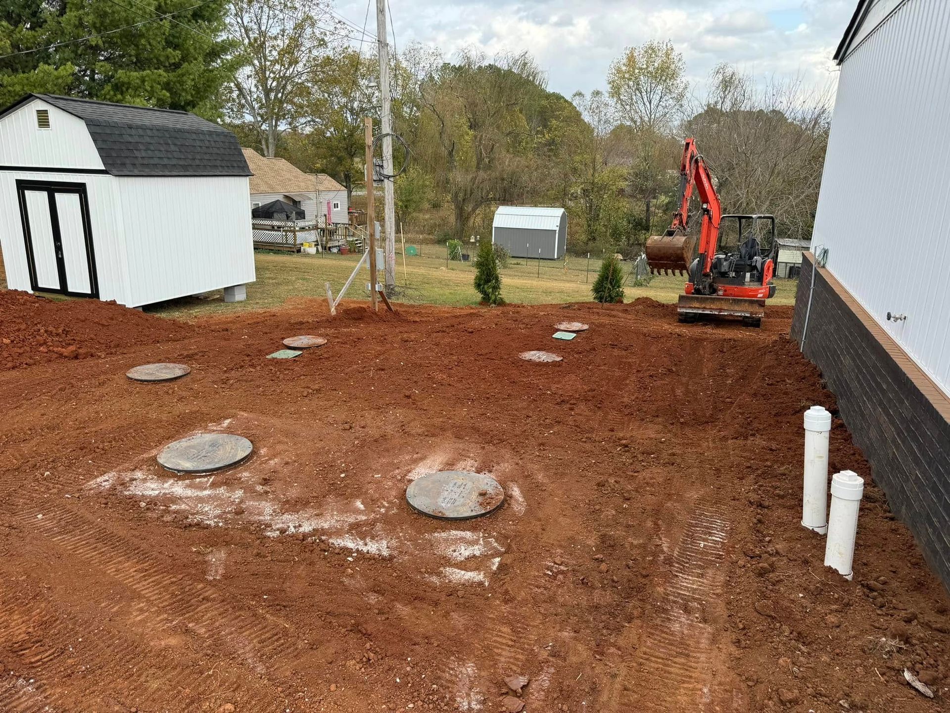 A red excavator is digging a hole in the dirt in front of a white shed.