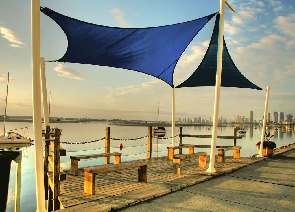 Two blue triangular shade sails over wooden benches on a waterfront pier with a city skyline in the distance at sunset.