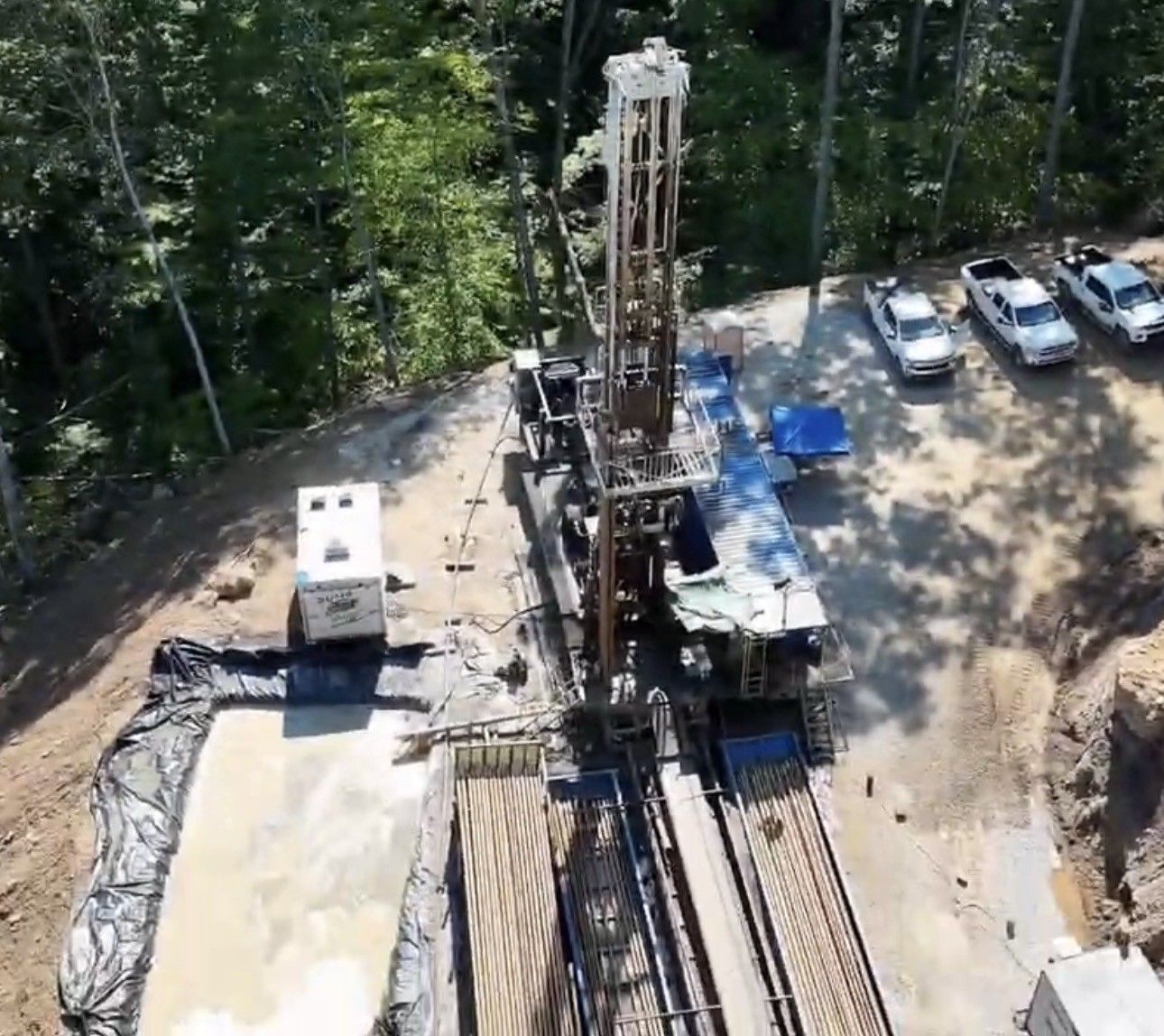 Aerial view of a drilling rig on a wooded construction site with parked vehicles and equipment nearby