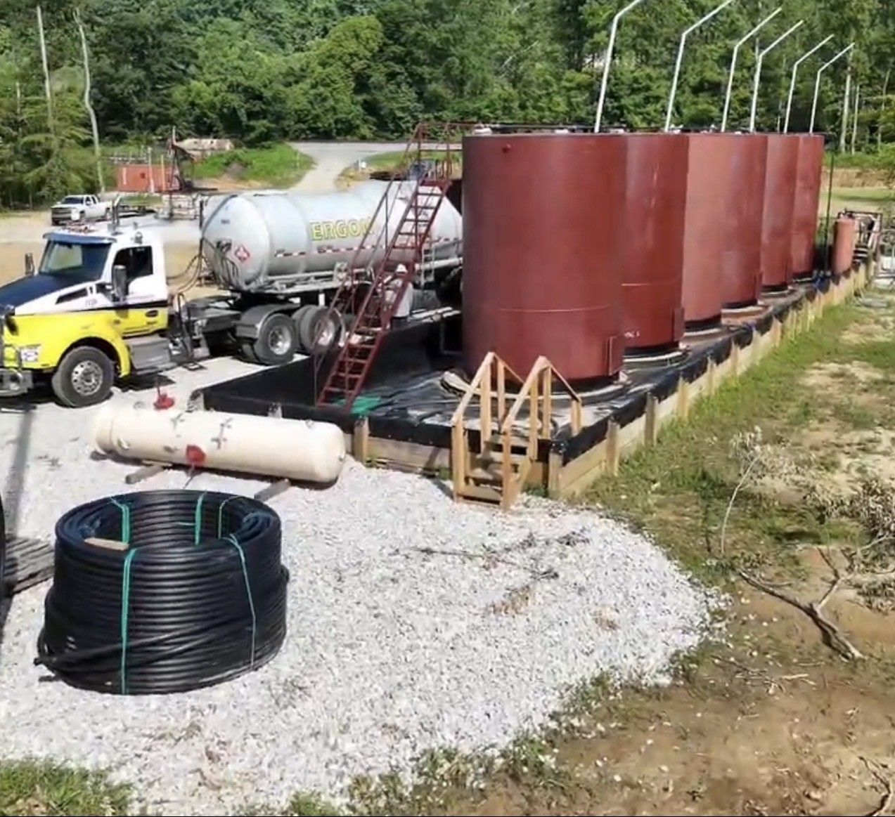 Industrial tanks and trucks at a roadside work site beside a gravel area and trees