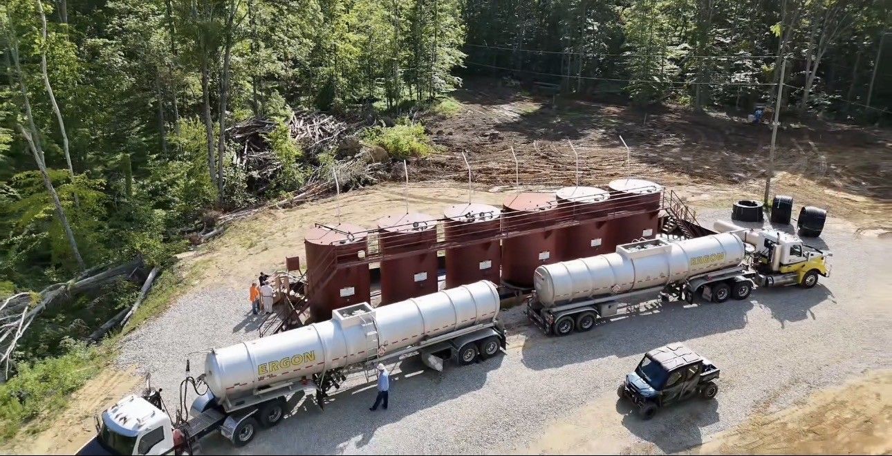 Overhead view of two white tanker trucks beside red railcars on a gravel lot near trees