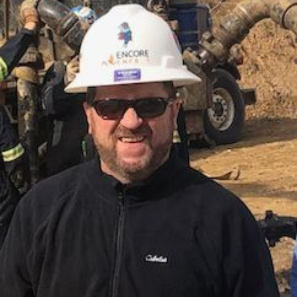 Worker in white hard hat and sunglasses smiling at a construction site with heavy equipment behind him