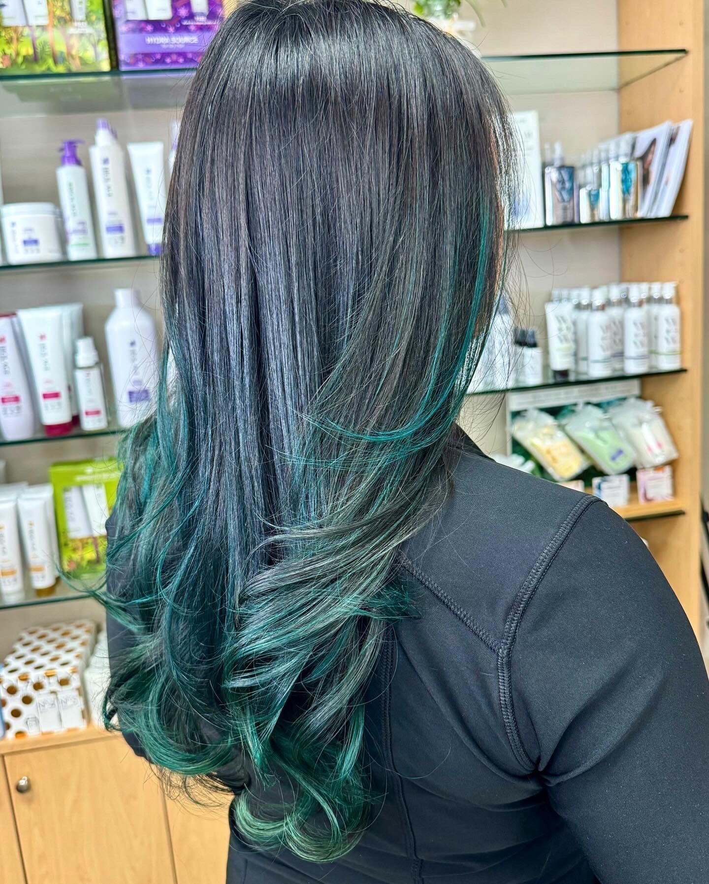 A woman with long black hair and green highlights is standing in front of a shelf of hair products.