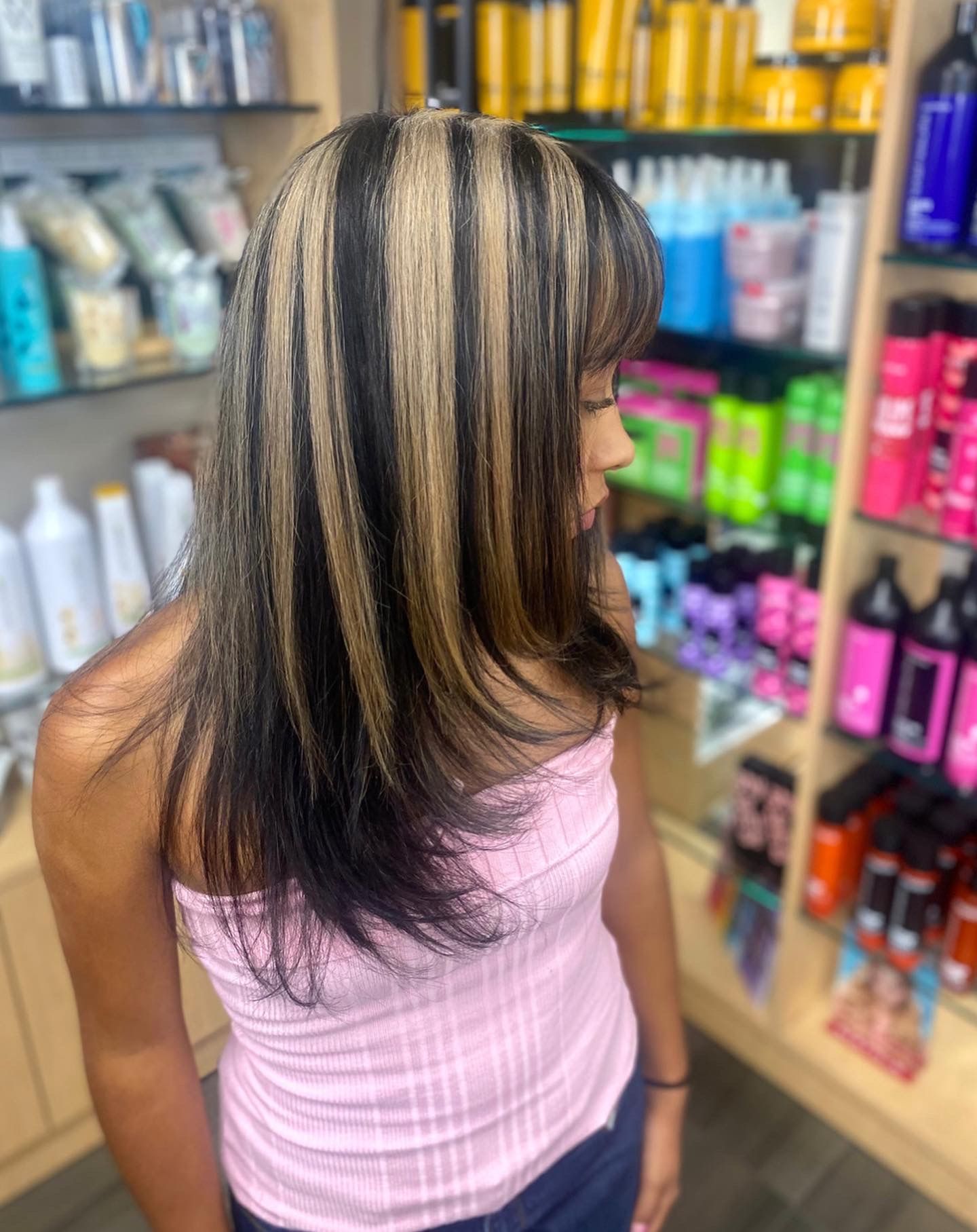 A woman with long black hair and blonde highlights is standing in a salon.