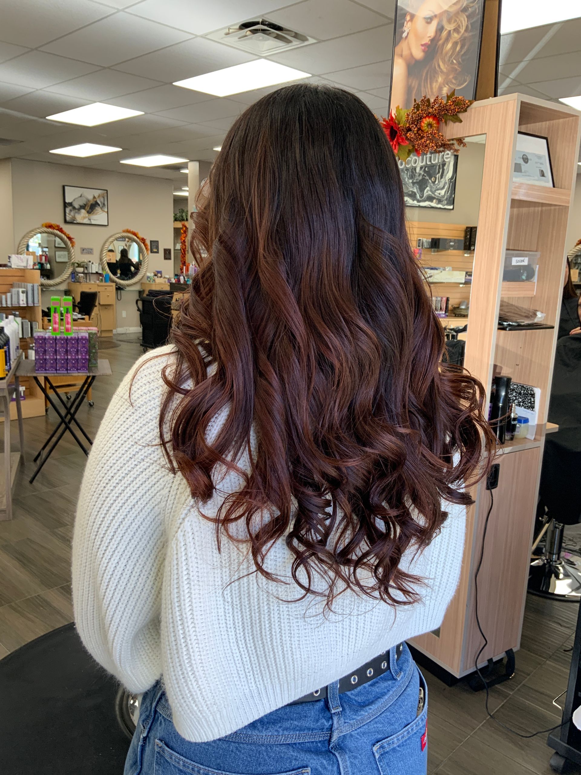 A woman with long brown hair is standing in a salon wearing a white sweater and jeans.