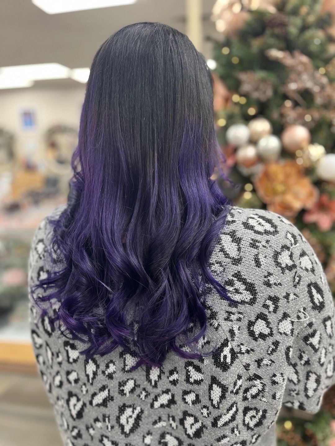 A woman with purple hair is wearing a leopard print sweater and standing in front of a christmas tree.