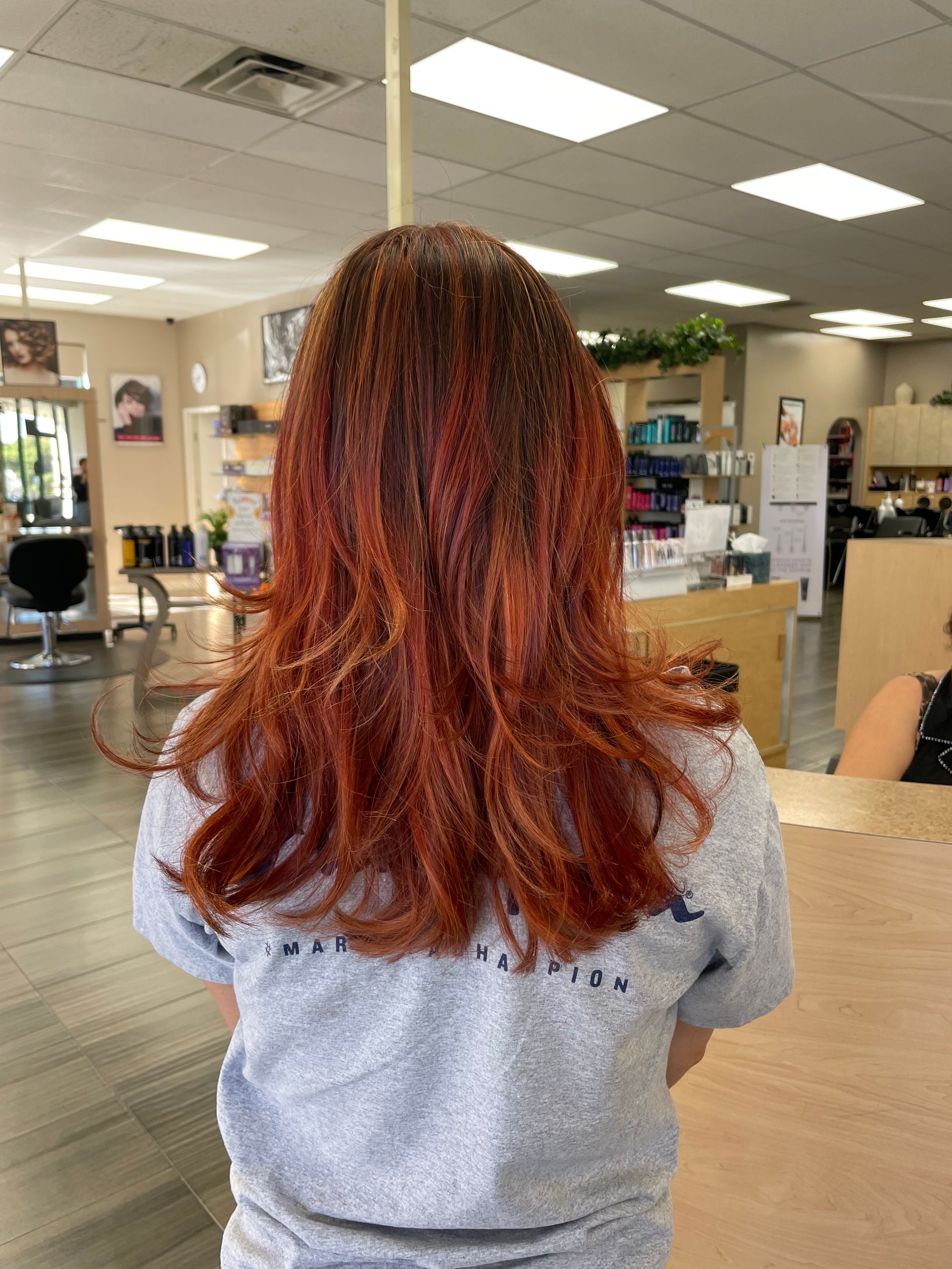 A woman with red hair is standing in a salon wearing a gray shirt.