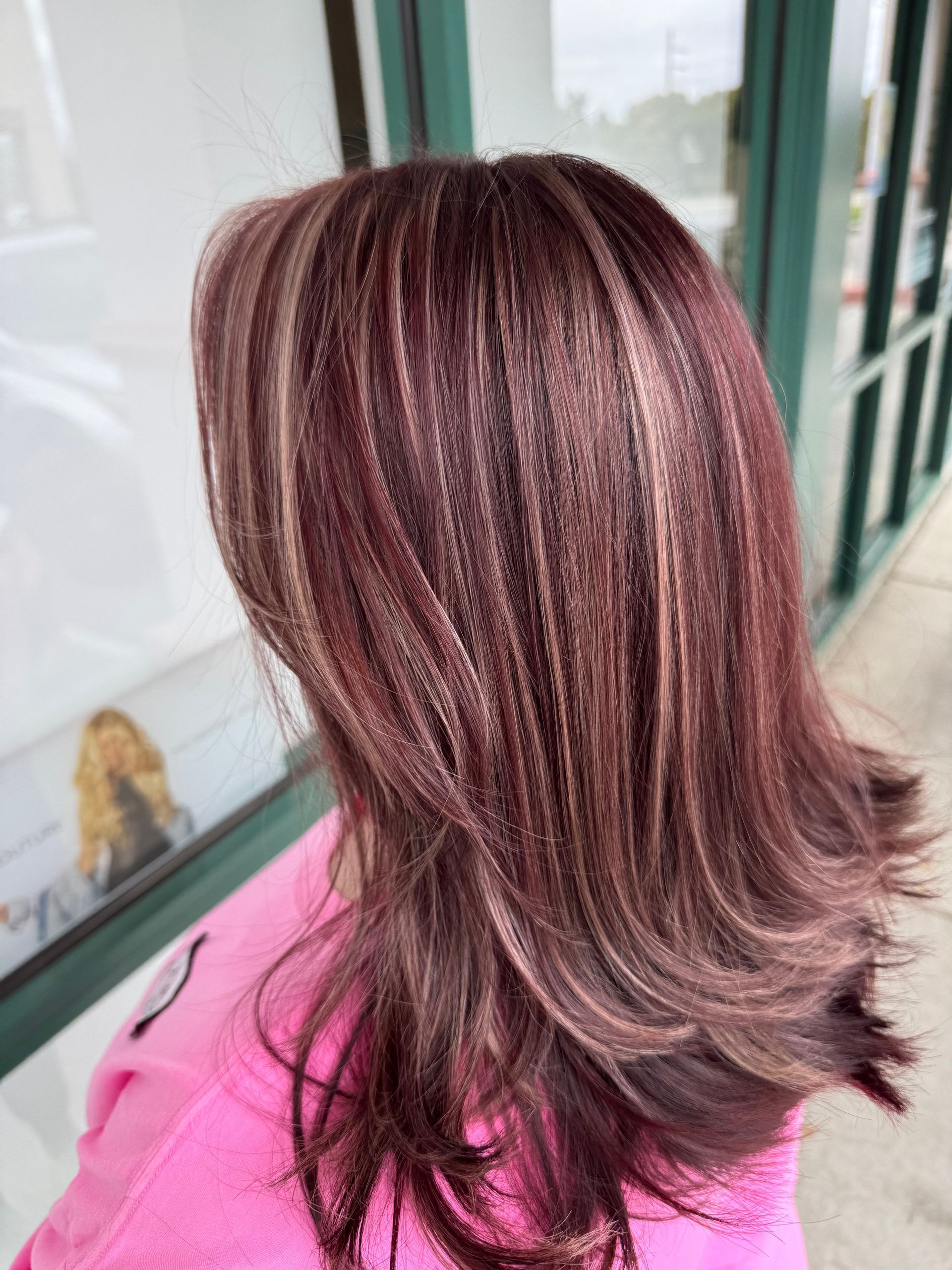 A woman with long brown hair is wearing a pink shirt and standing in front of a window.