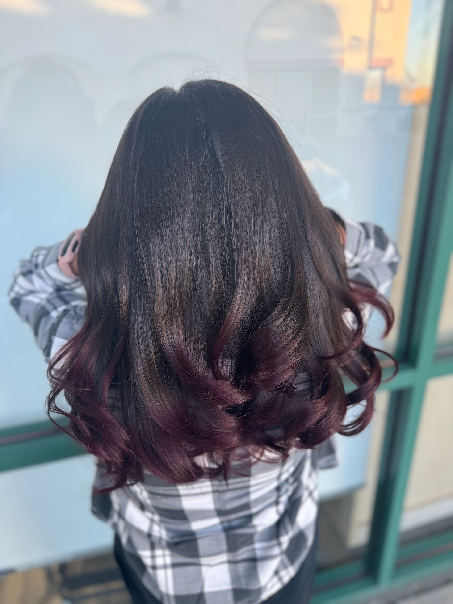 A woman wearing a plaid shirt and burgundy hair is standing in front of a window.
