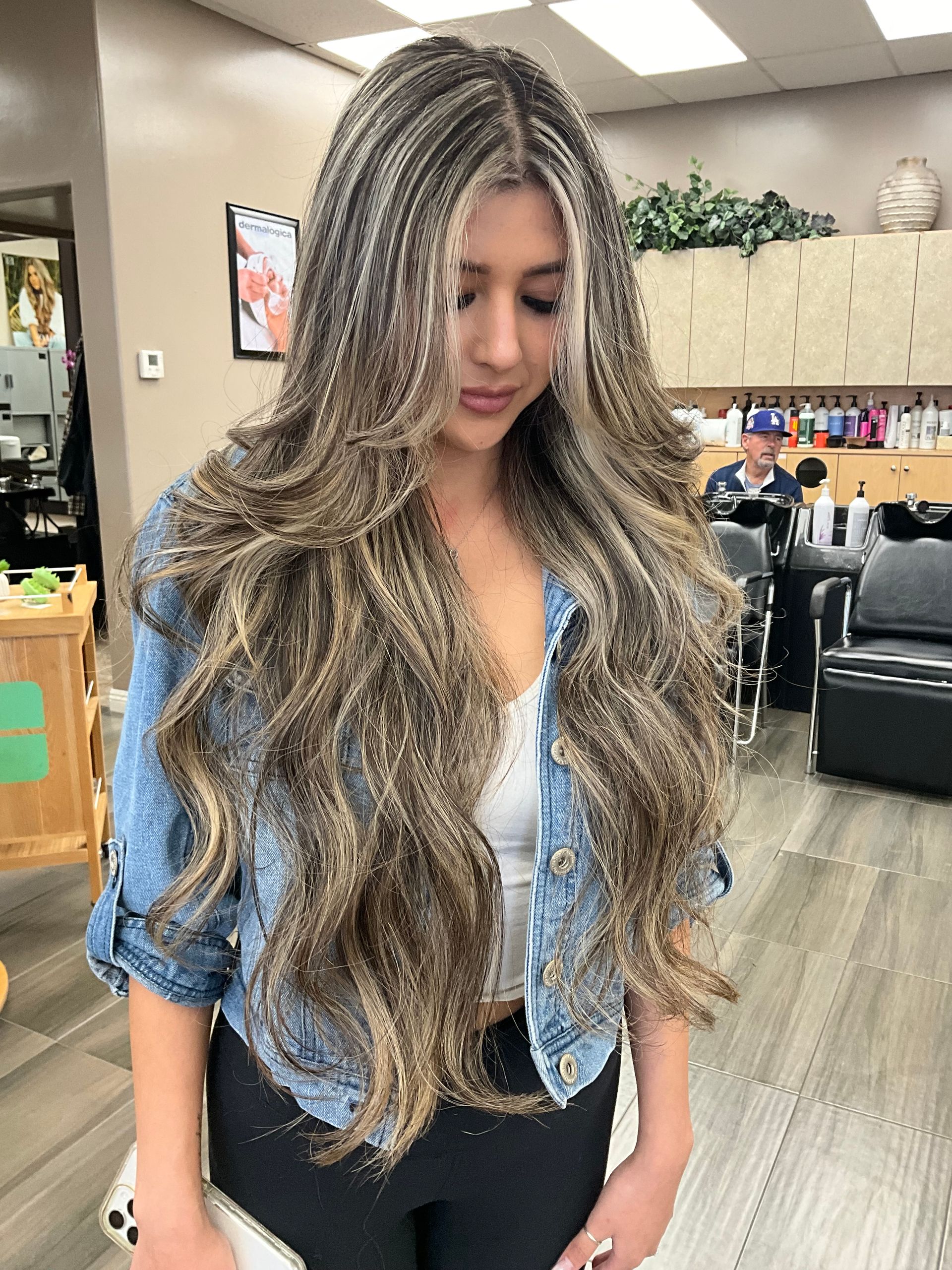 A woman with long hair is standing in a salon wearing a denim jacket.
