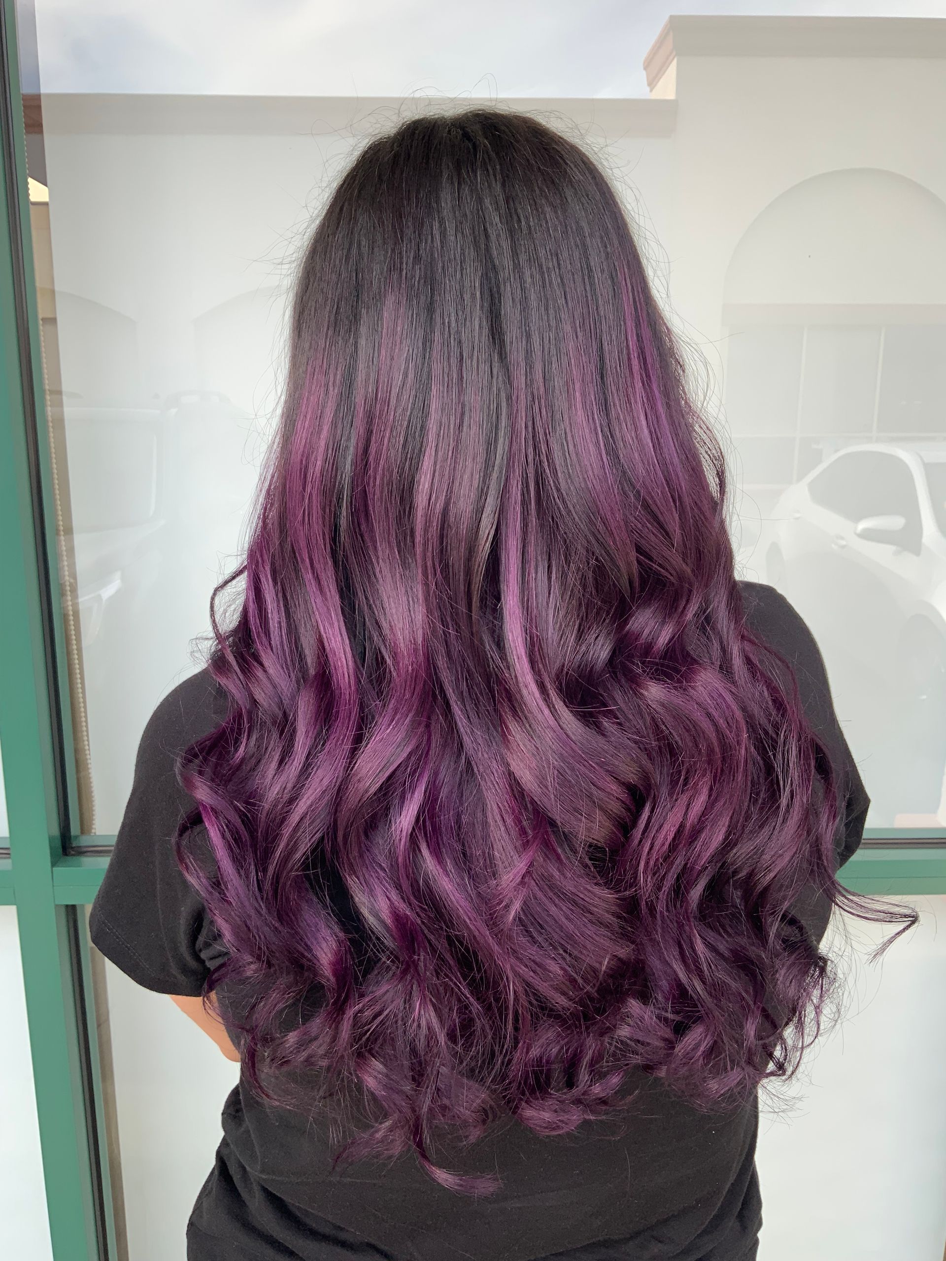 A woman with long purple hair is standing in front of a window.