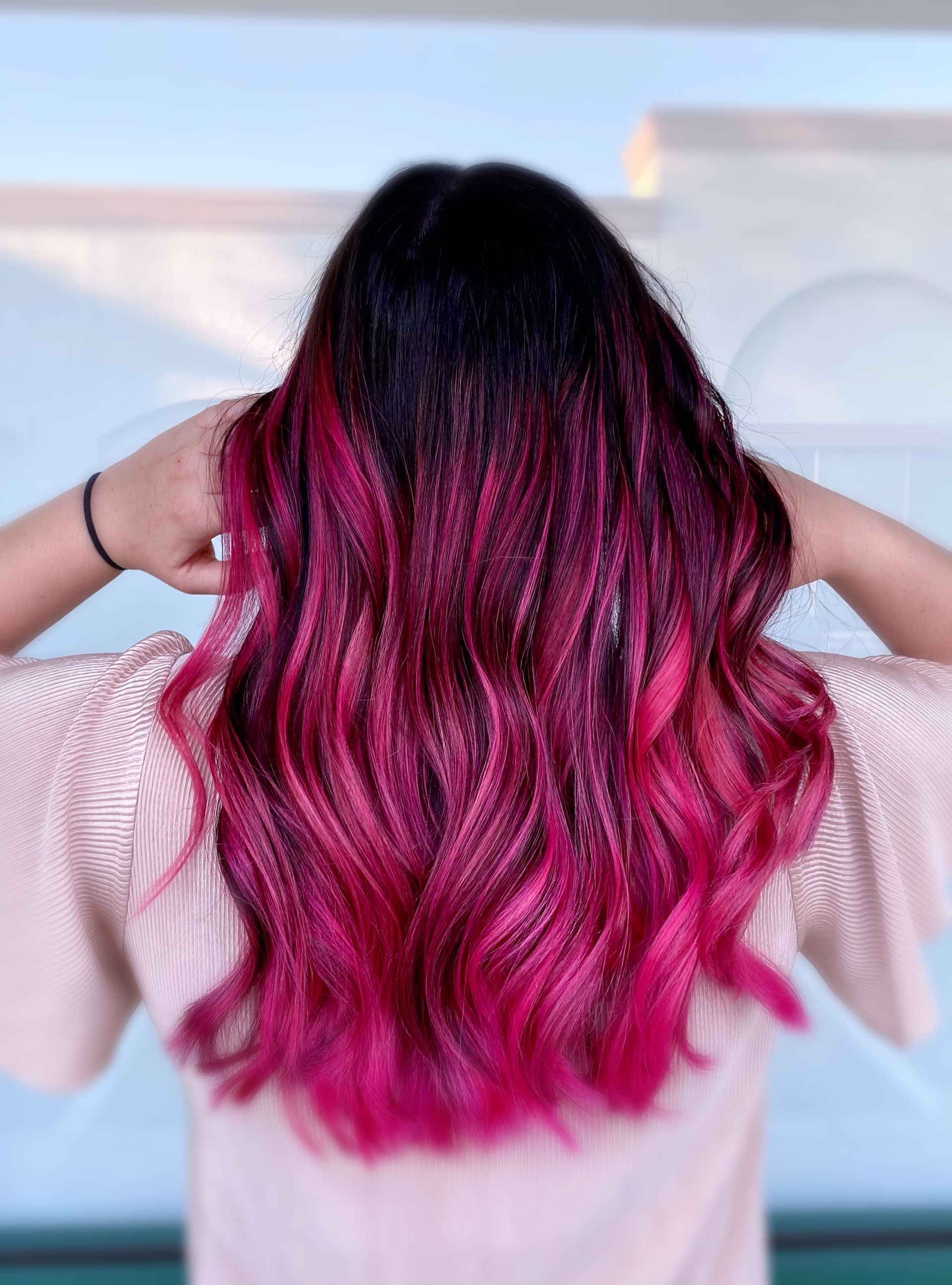 A woman with long pink hair is standing in front of a window.