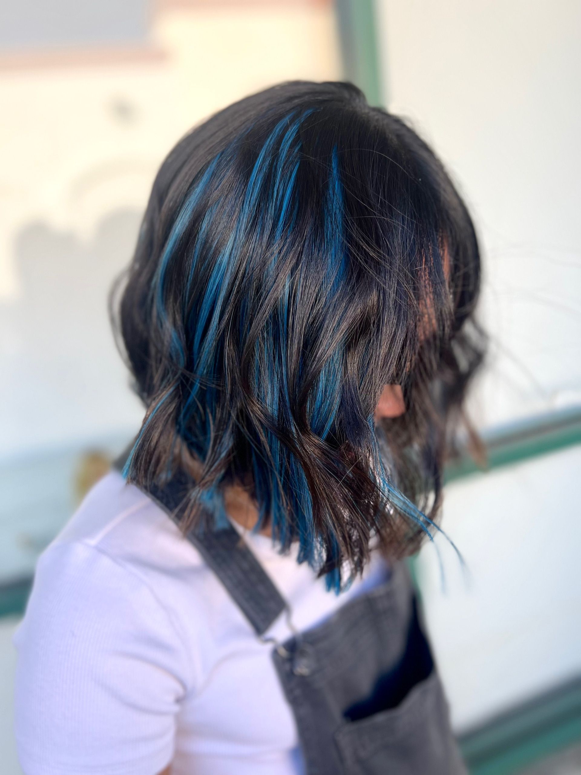 A woman with black hair and blue highlights is wearing overalls and a white shirt.