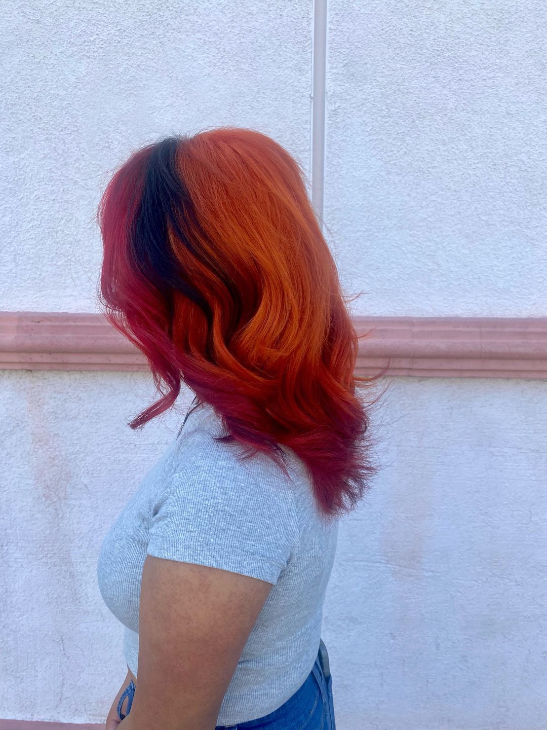 A woman with red and black hair is standing in front of a white wall.