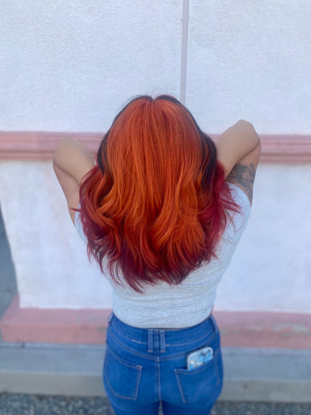 A woman with red hair is standing in front of a wall with her hands behind her head.