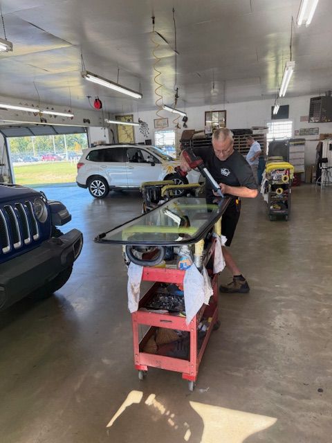 Man applies sealant to car window on a cart in a repair shop. A Jeep and SUV are also present.