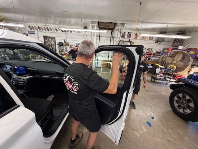 A man installs a car window in a garage. Another person stands nearby.