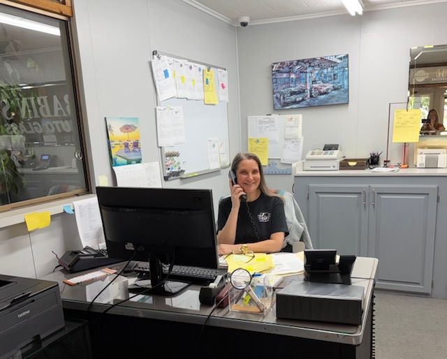 Woman at desk talking on phone, smiling. Office setting with computer, printer, and whiteboard.