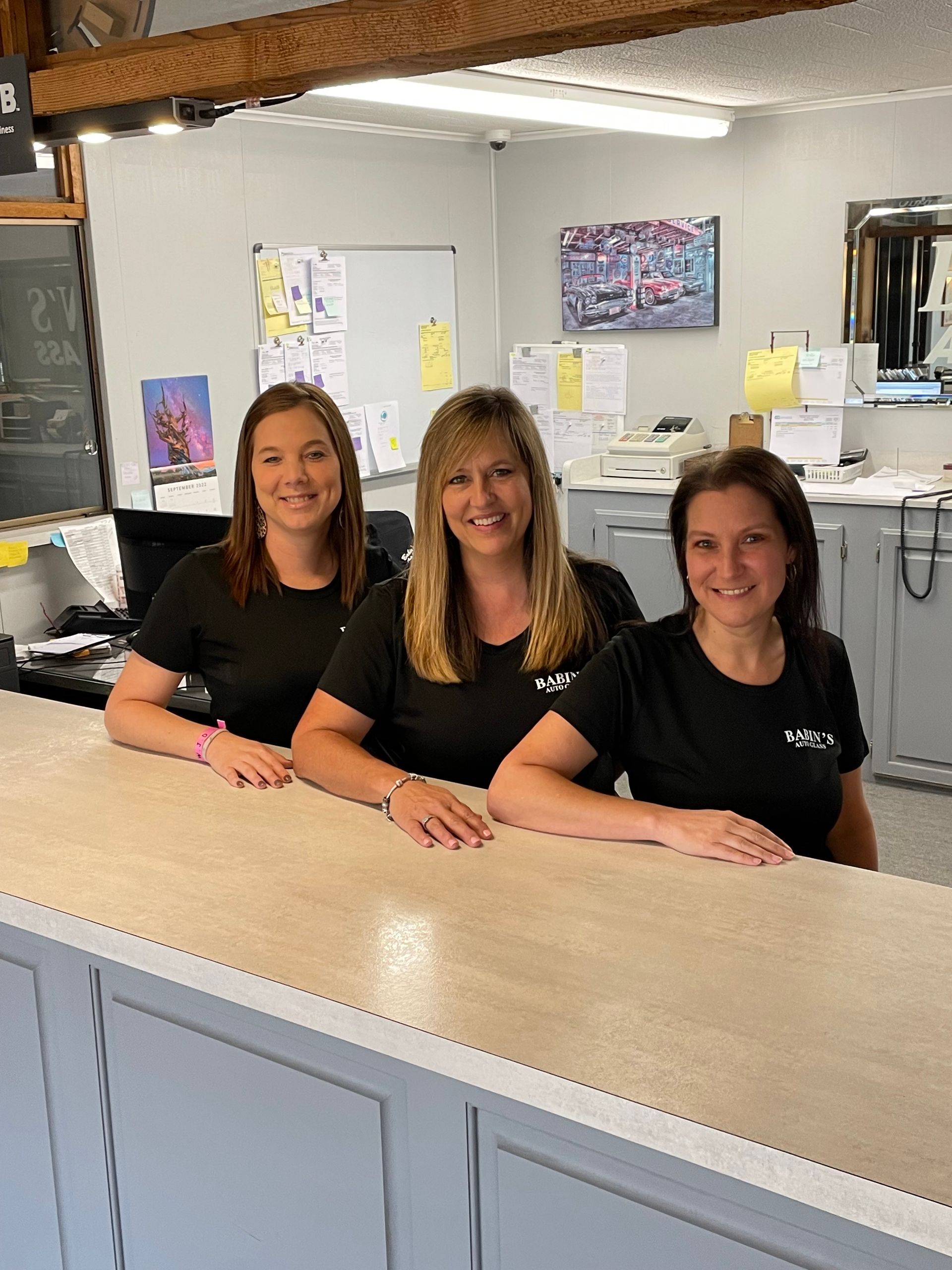 Three women smiling behind a counter in an office setting, wearing black shirts.