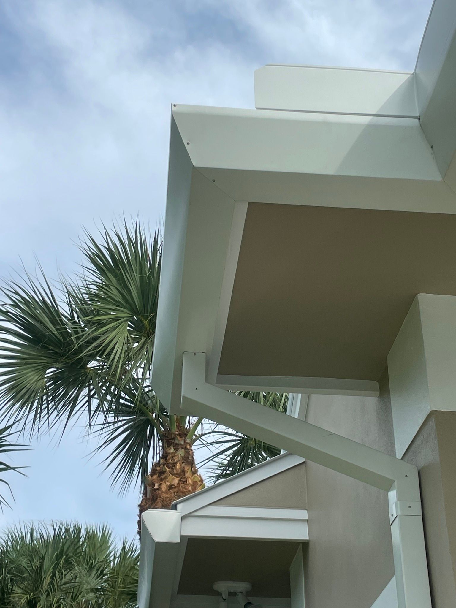 Exterior corner of a building with white trim and beige walls, palm tree in the background, against a cloudy sky.