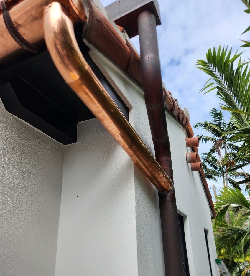 Copper gutters and downspout on a white stucco building, angled view with a blue sky and palm trees in the background.