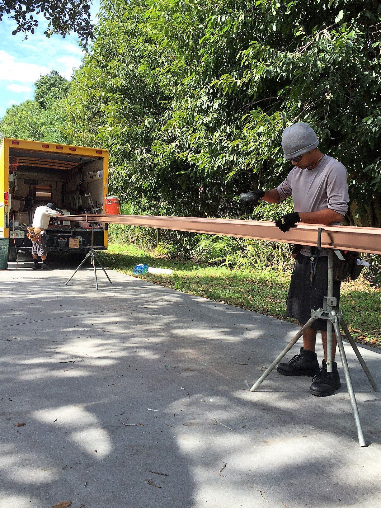 A person wearing gloves and a hard hat is sanding a long piece of wood on a sawhorse, next to a yellow truck, outdoors.