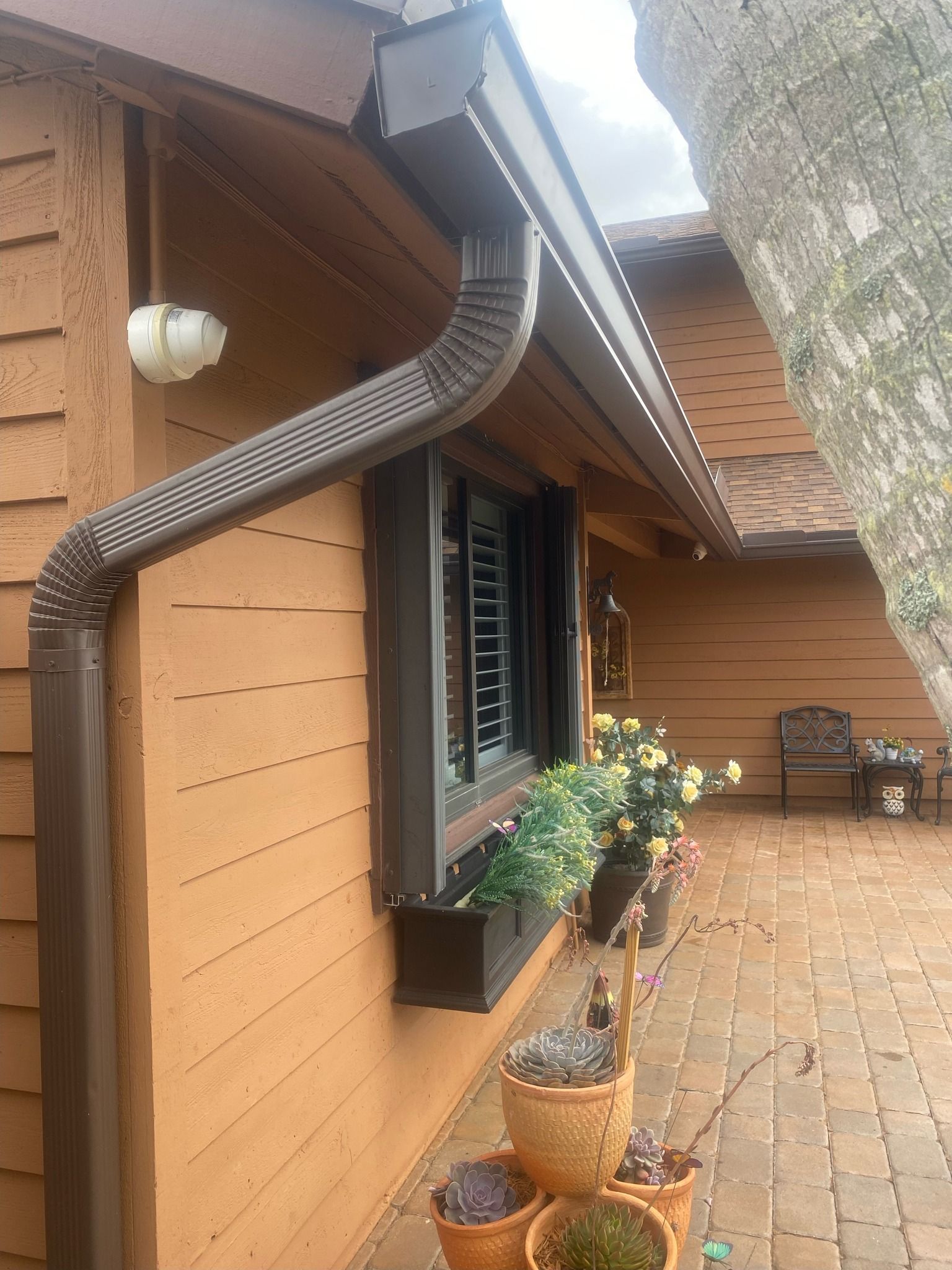 Brown gutter and downspout on a brown house with a window box and potted plants.