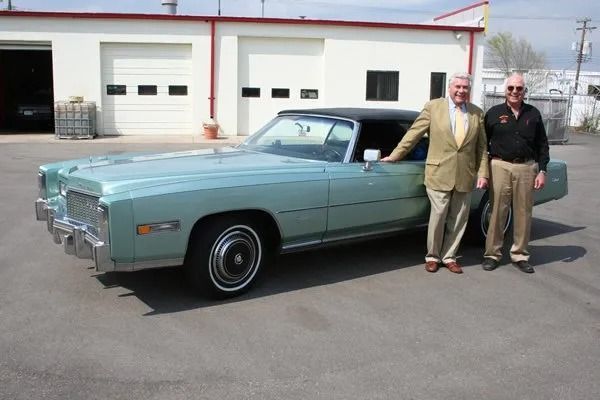Two men stand beside a light-green Cadillac convertible in front of a building with garage doors.