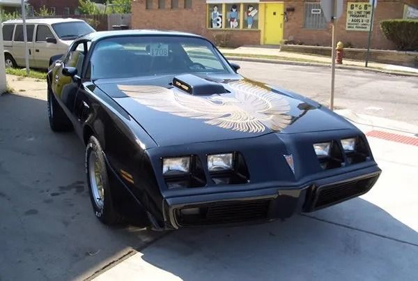 Black Pontiac Trans Am with a gold eagle emblem on the hood, parked on a city street.