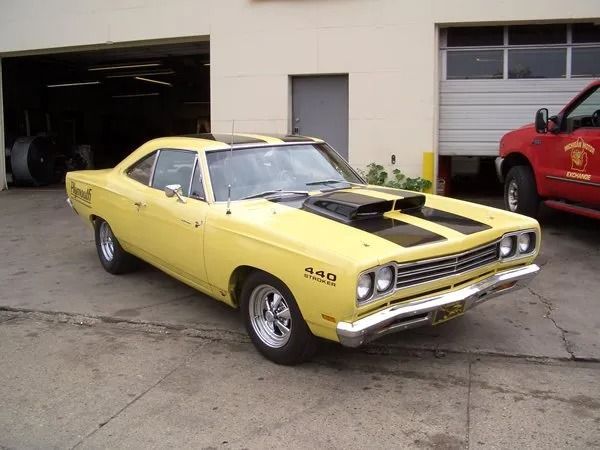 Yellow classic Plymouth Road Runner car with black stripes and hood scoop parked in front of a garage.