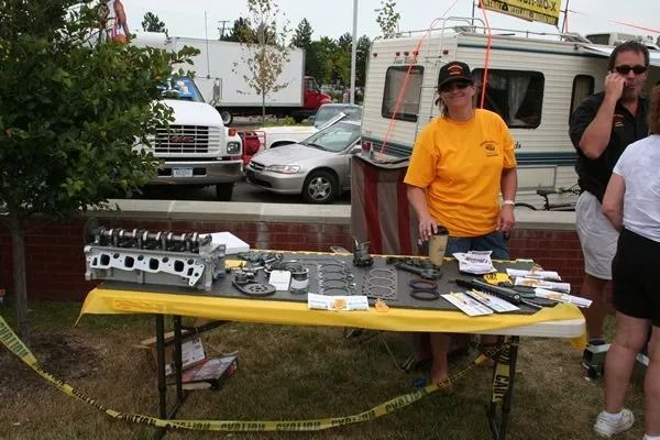 A woman stands at a table displaying car parts; outdoors event setting.