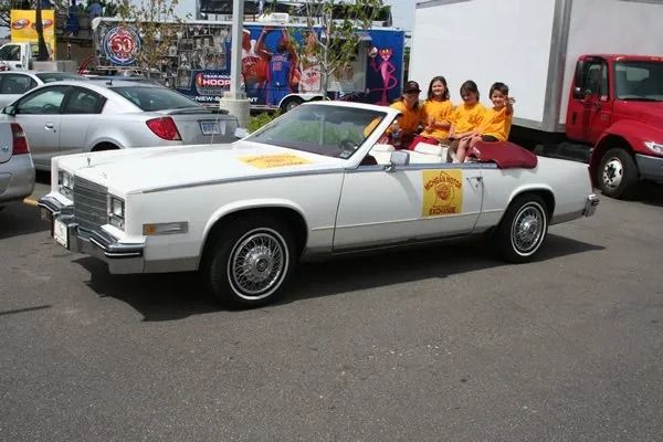 White convertible car with people in orange shirts, parked, sunny day.