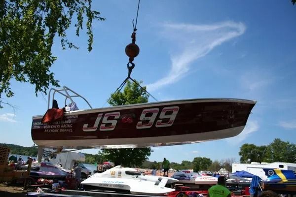 Race boat JS 99 lifted by a crane at an outdoor event, with other boats and people visible.