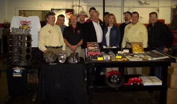 Group of people with car engine parts laid out on a table in a garage.
