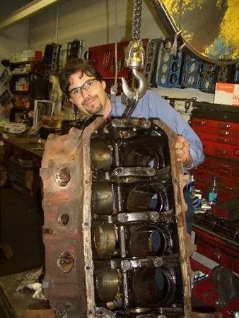 Man holding a large engine block in a garage, smiling. The block is suspended by a hook.