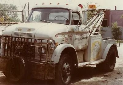 White, weathered tow truck, possibly 1960s era, with a light on top and a tire hanging on the front.