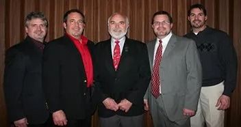 Five men in suits and casual wear stand in front of a curtain.