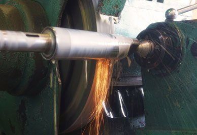 A cylindrical metal part being ground by a rotating abrasive wheel, creating sparks, in a machine shop.