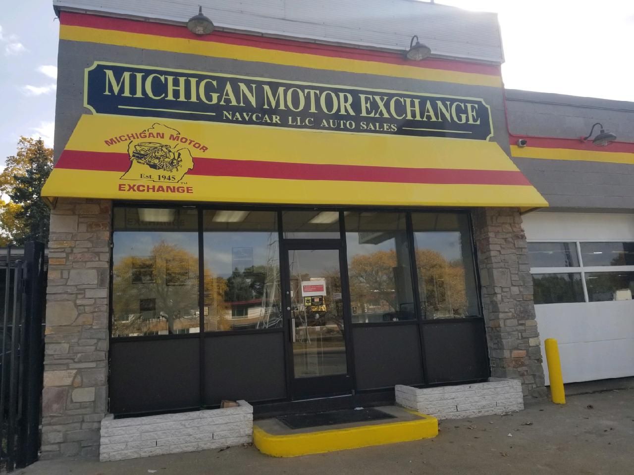 Exterior of Michigan Motor Exchange car dealership. Yellow and red awning, stone accents, glass windows, and a closed door.