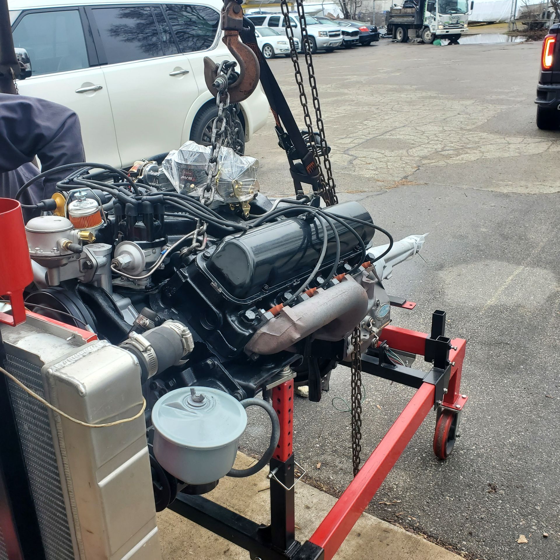 Engine on a stand being lifted with a crane hook; outdoors, asphalt ground, other vehicles in background.