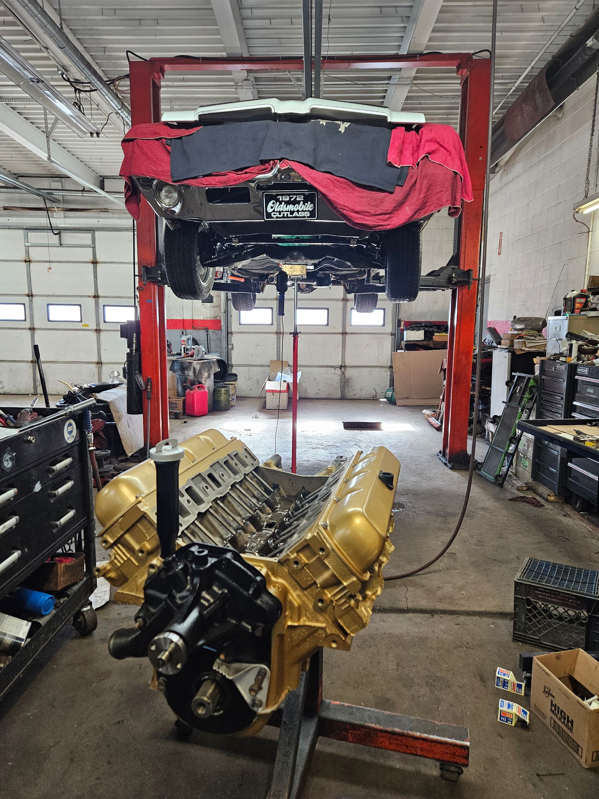 Engine repair shop: car on lift, golden engine in foreground, tools and parts visible.