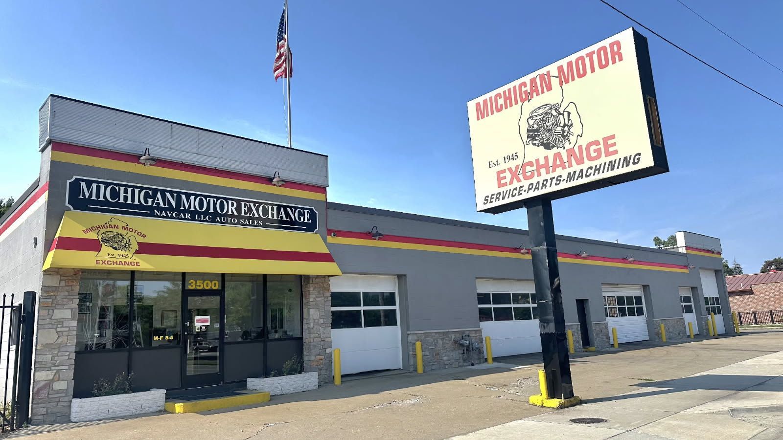 Michigan Motor Exchange, auto shop with American flag and sign. Gray building with garage doors.