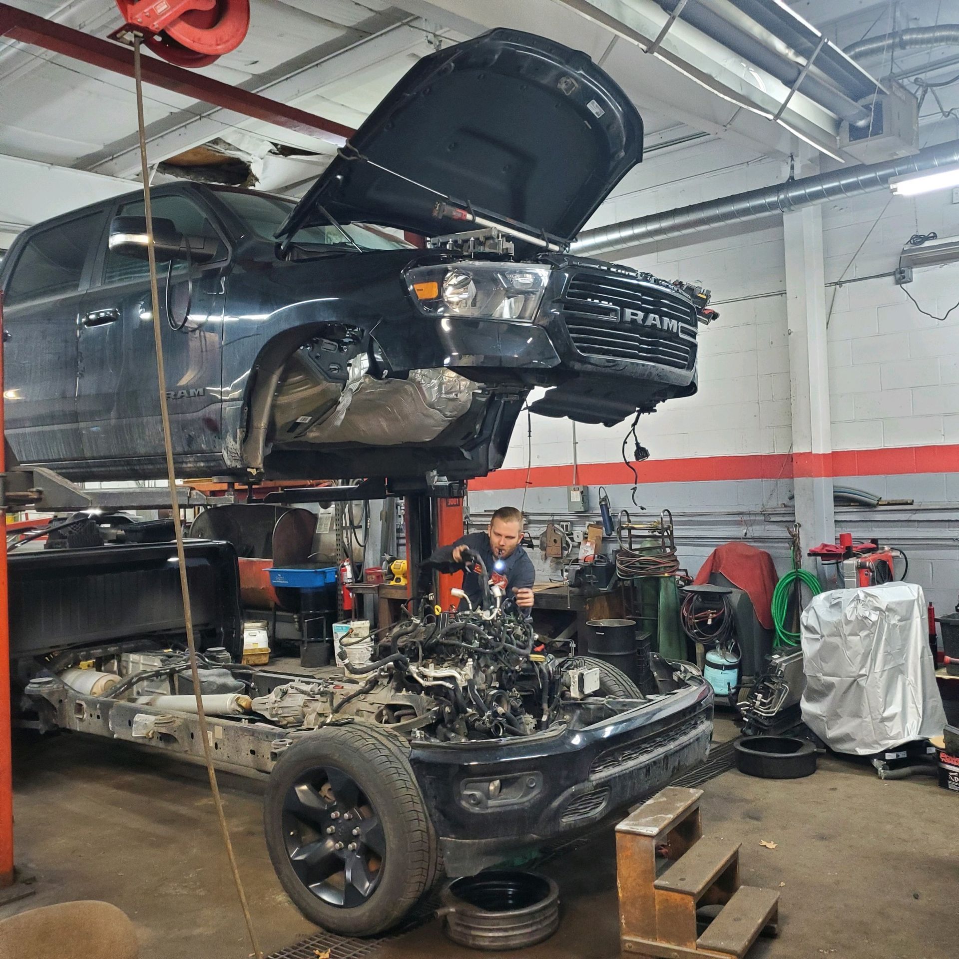 Black pickup truck being repaired in a garage, lifted on a hoist with the hood and front end removed, a mechanic working.