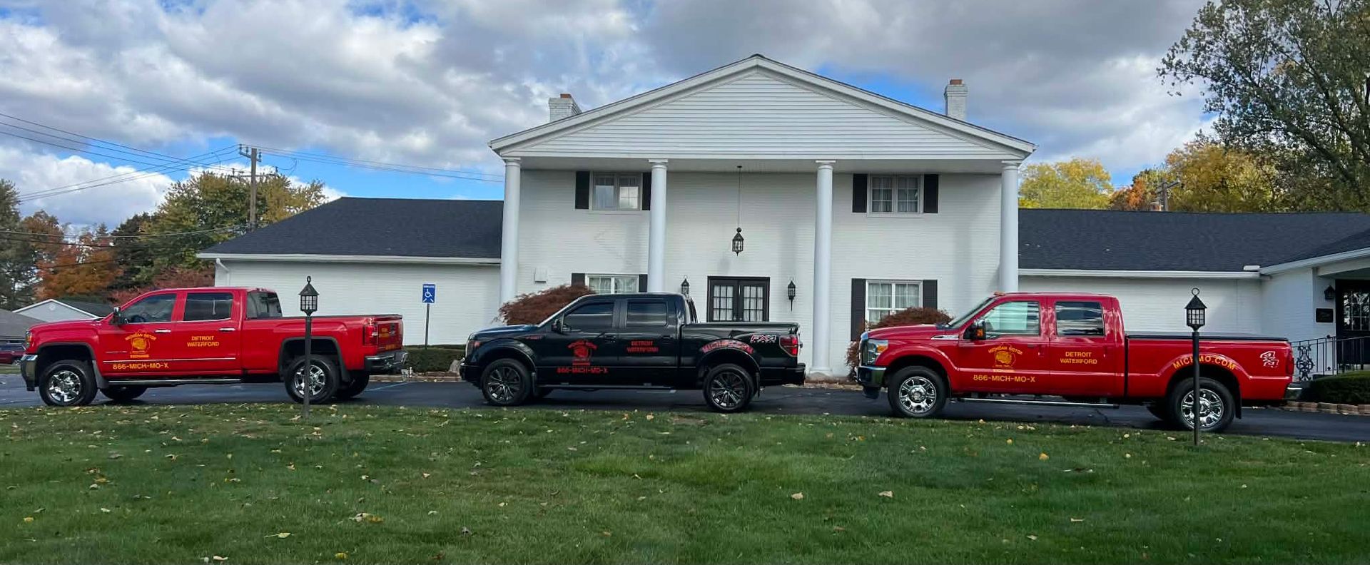Three lifted red and black pickup trucks parked in front of a white building with pillars. Cloudy sky.