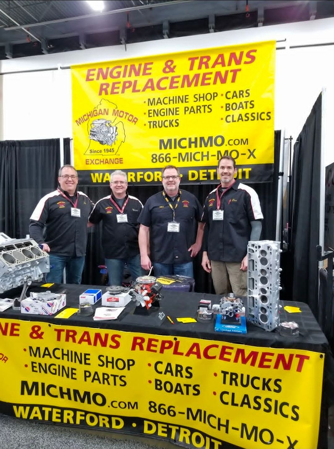 Four men at a booth promoting engine and transmission replacement services at a tradeshow.