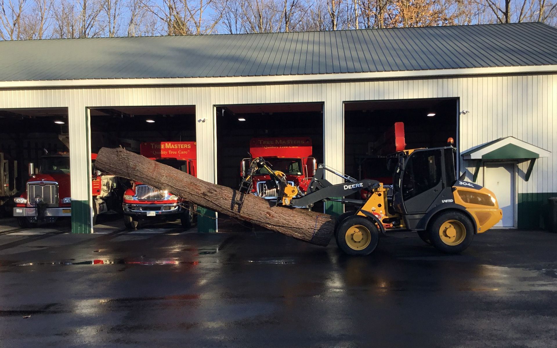 A bulldozer is carrying a large log in front of a fire station.