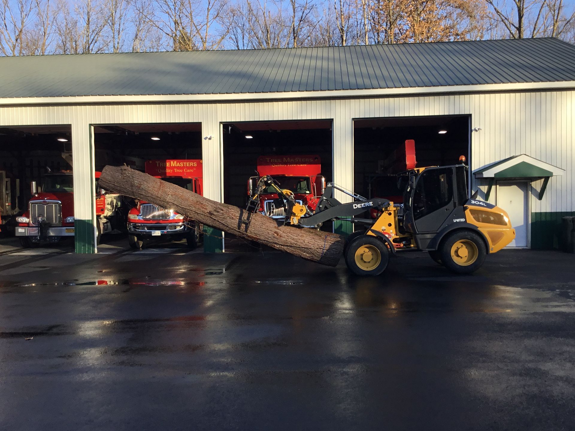 A yellow tractor is carrying a large log in front of a garage